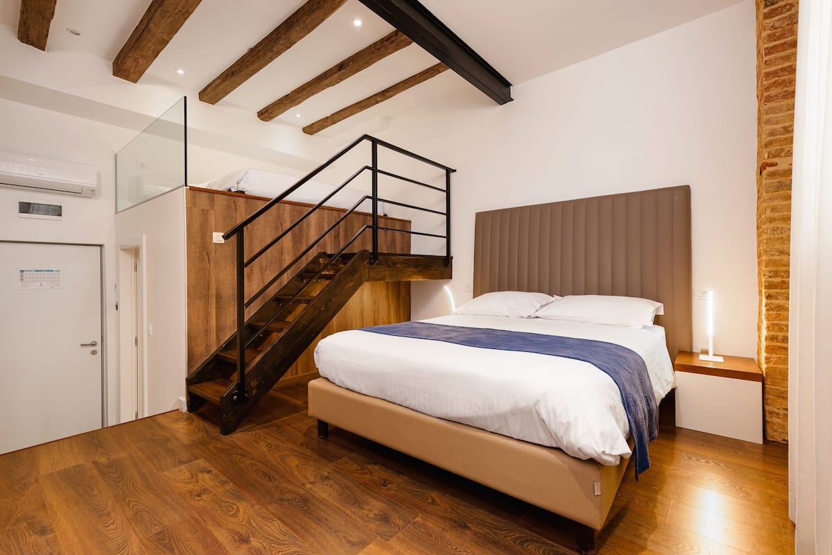 Un dormitorio con una escalera de madera, una cama con cabecera, una mesita de noche con una lámpara y un suelo de madera.