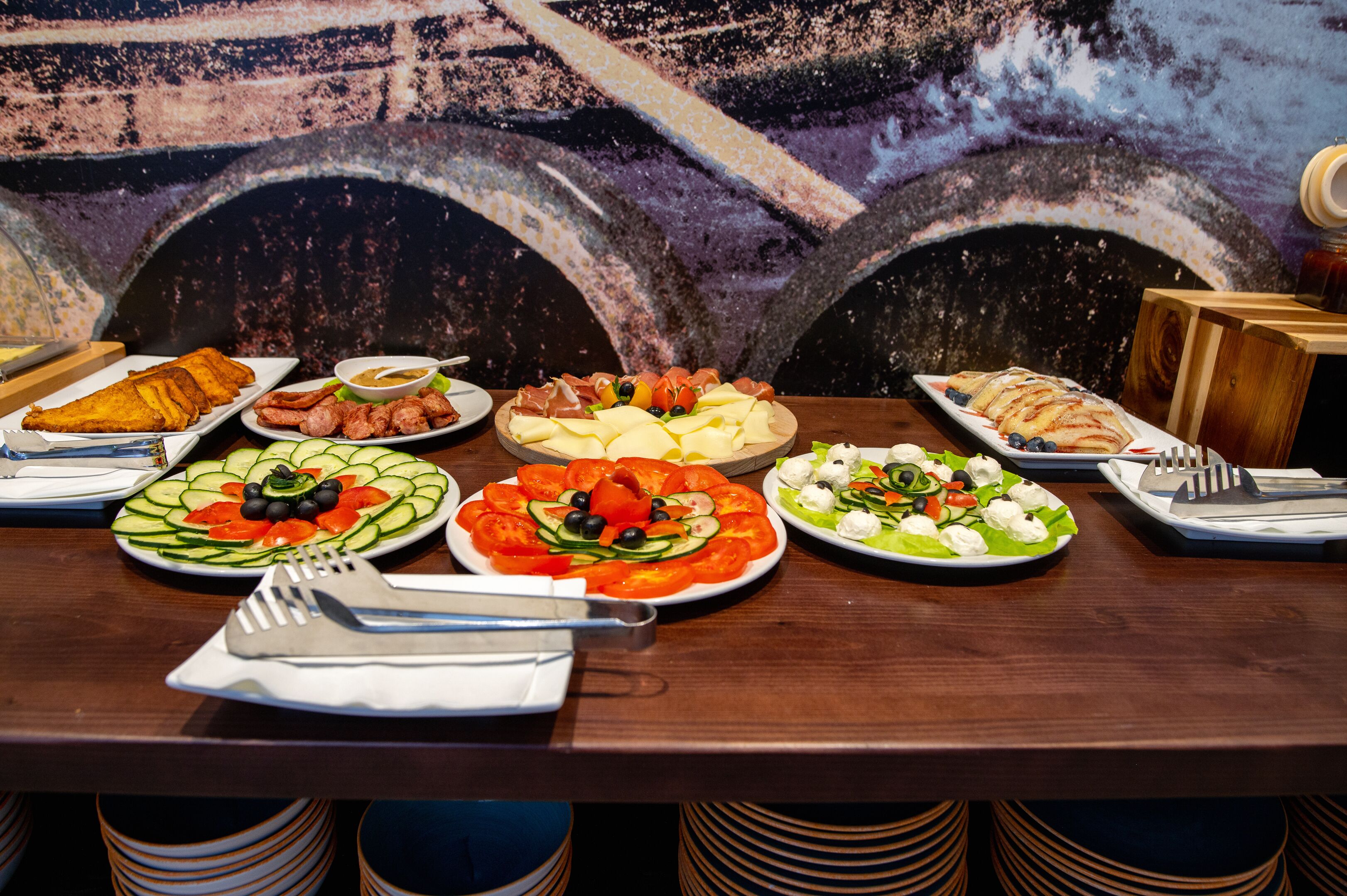 Une table dressée avec divers plats, notamment des salades, des viandes et des pâtisseries, le tout mis en valeur par une fresque murale en arrière-plan.