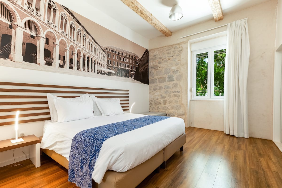 Un dormitorio con cama, mesita de noche, pared de piedra y ventana con cortinas.