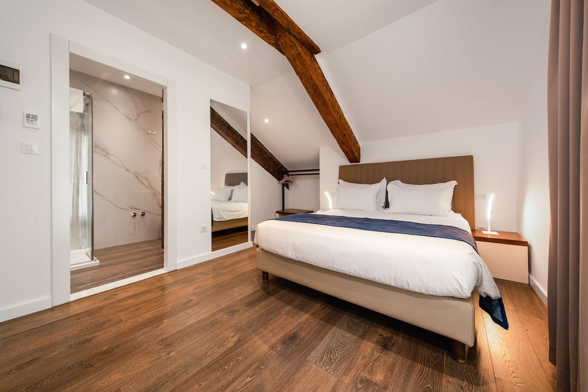 Un dormitorio moderno con una cama grande, vigas de madera, un vestidor y una ducha con mampara de vidrio.