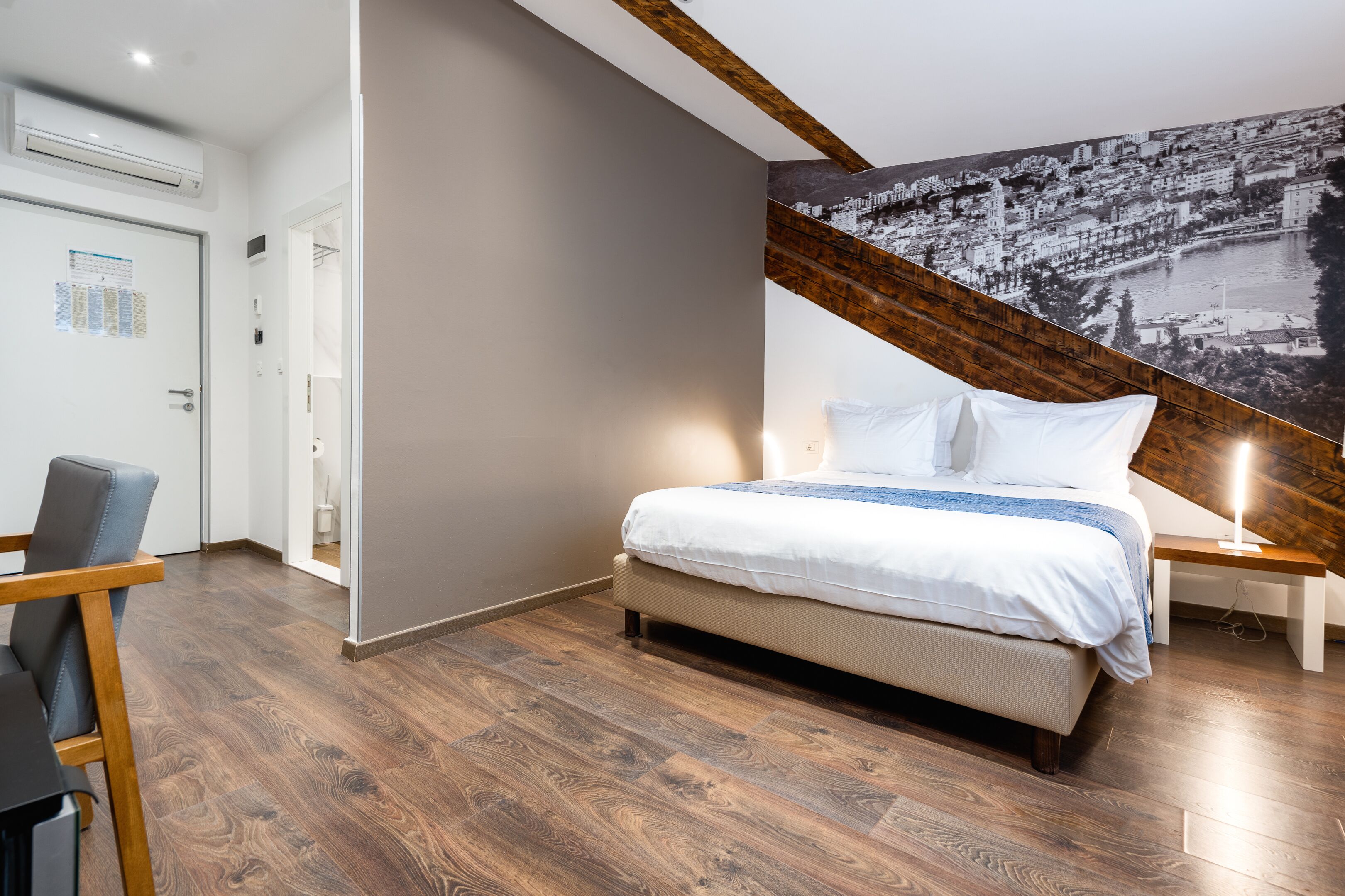 Un dormitorio moderno con suelo de madera, una cama con ropa blanca, un escritorio con silla y un mural de paisaje urbano en la pared.