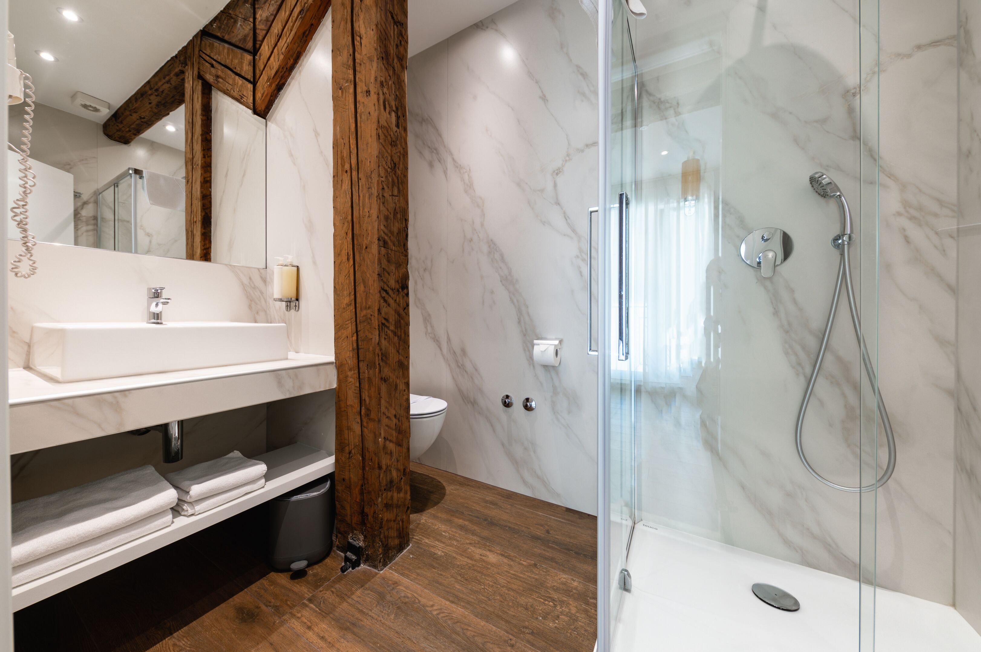 Un baño moderno con lavamanos de mármol, vigas de madera y cabina de ducha con paredes de vidrio.