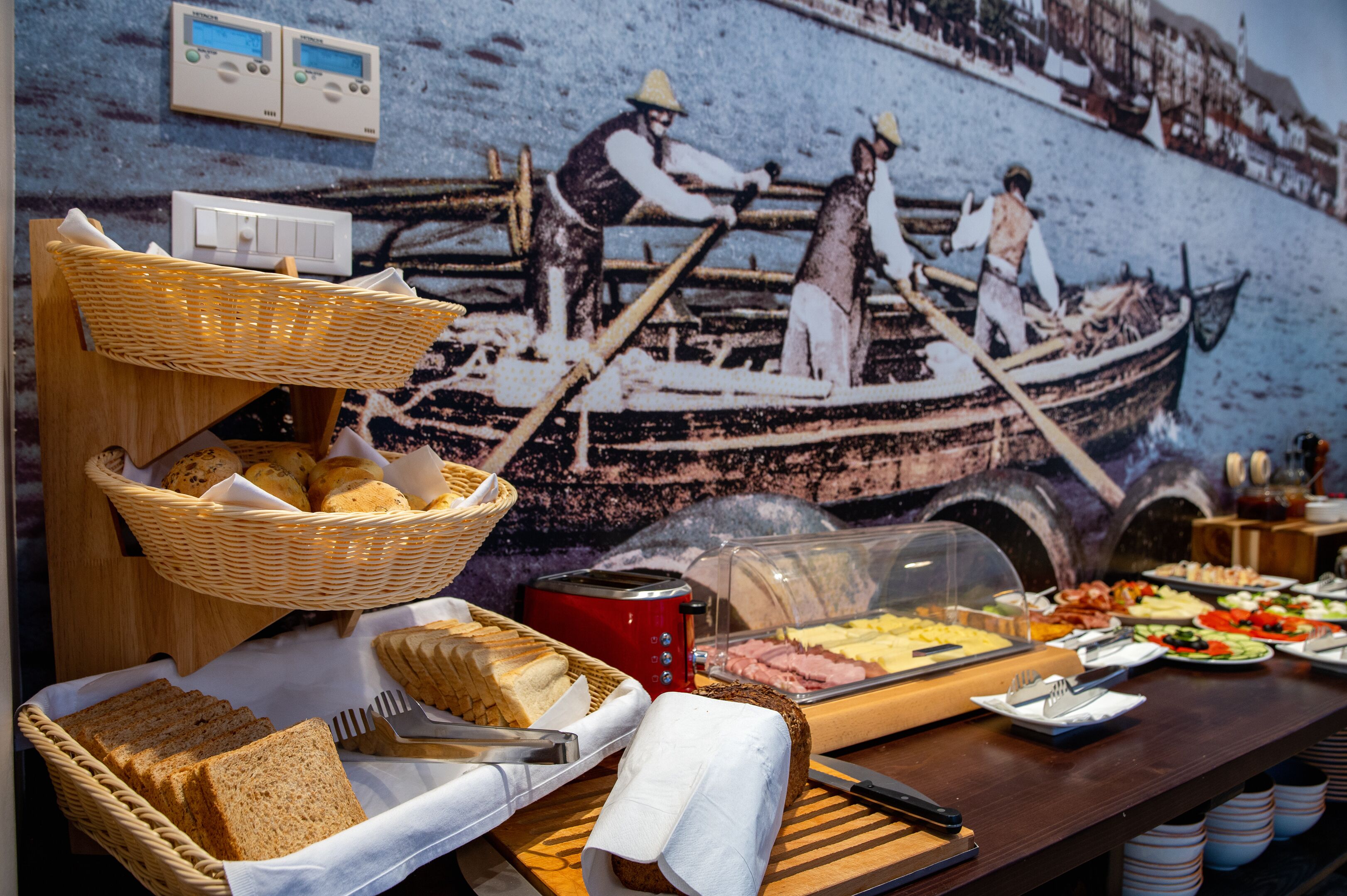 A buffet with various food items, baskets of bread, and a wall mural depicting a historical scene.