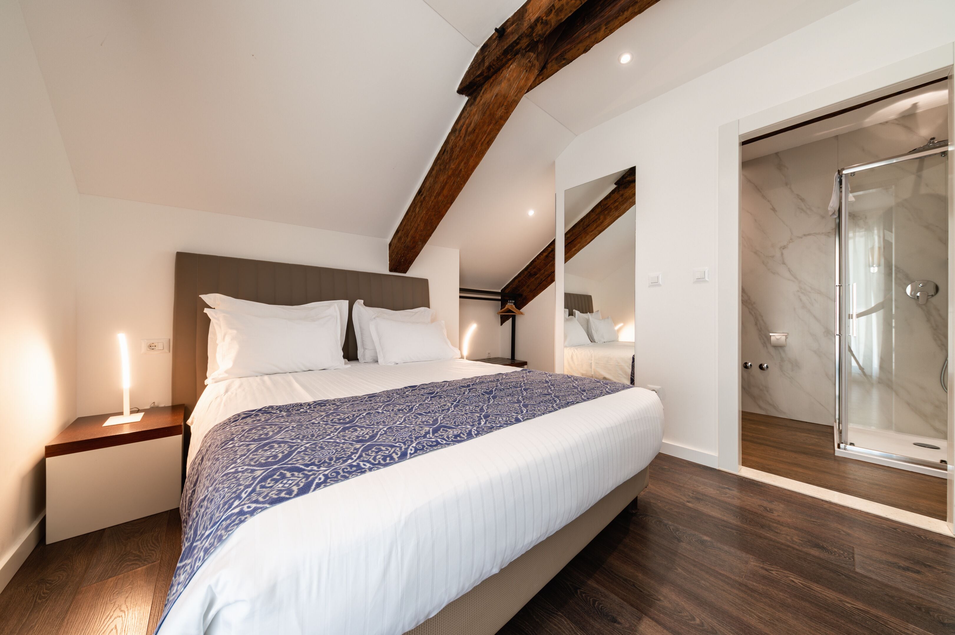 A bedroom with a large bed, wooden beams, a glass shower, and a marble floor.