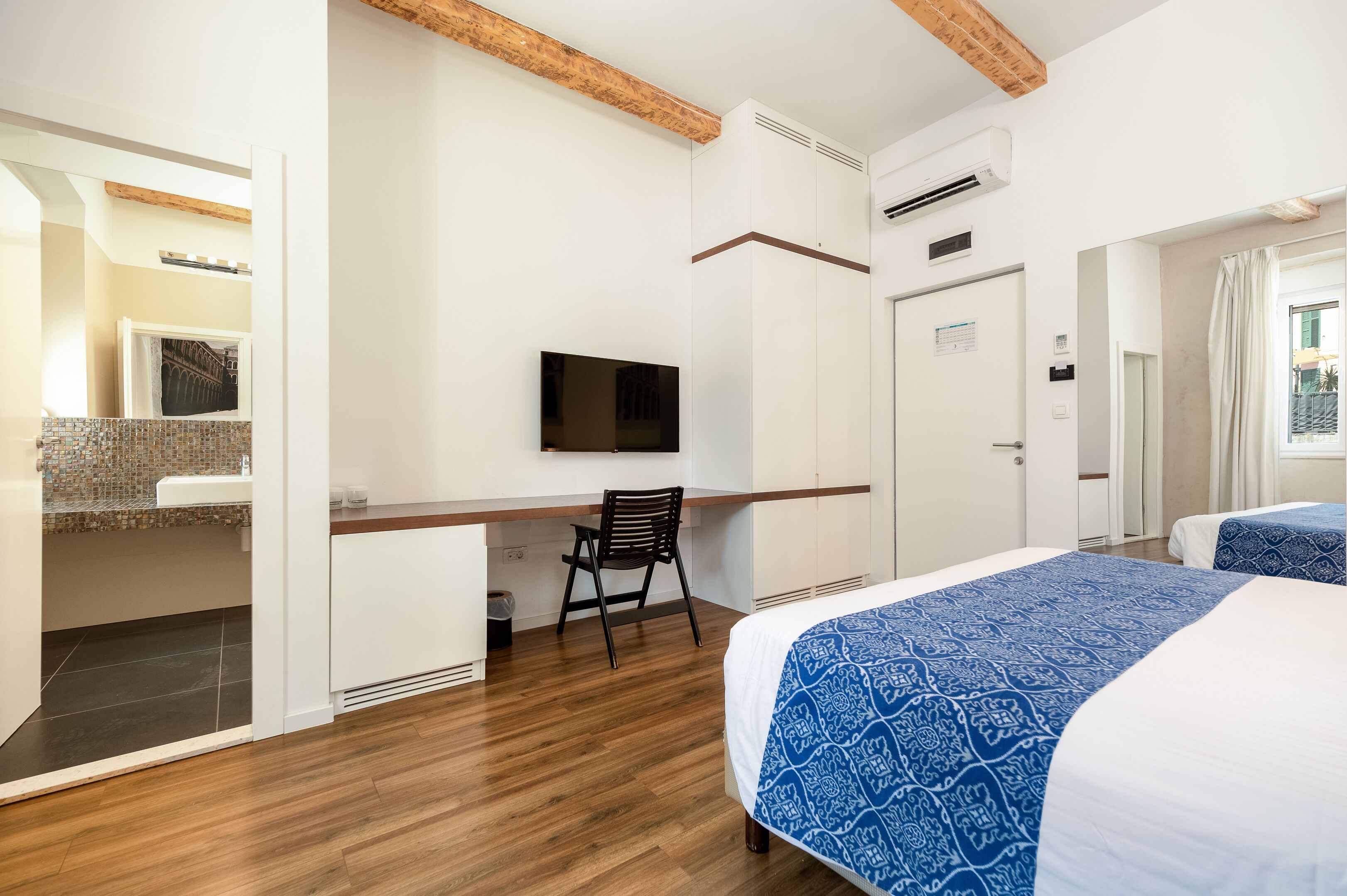 Een hotelkamer met een houten vloer, een bureau met stoel, een bed met blauw beddengoed en een aan de muur bevestigde televisie.