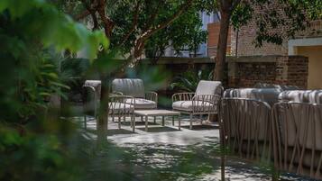 An outdoor seating area with cushioned chairs and tables, surrounded by trees and a brick wall.