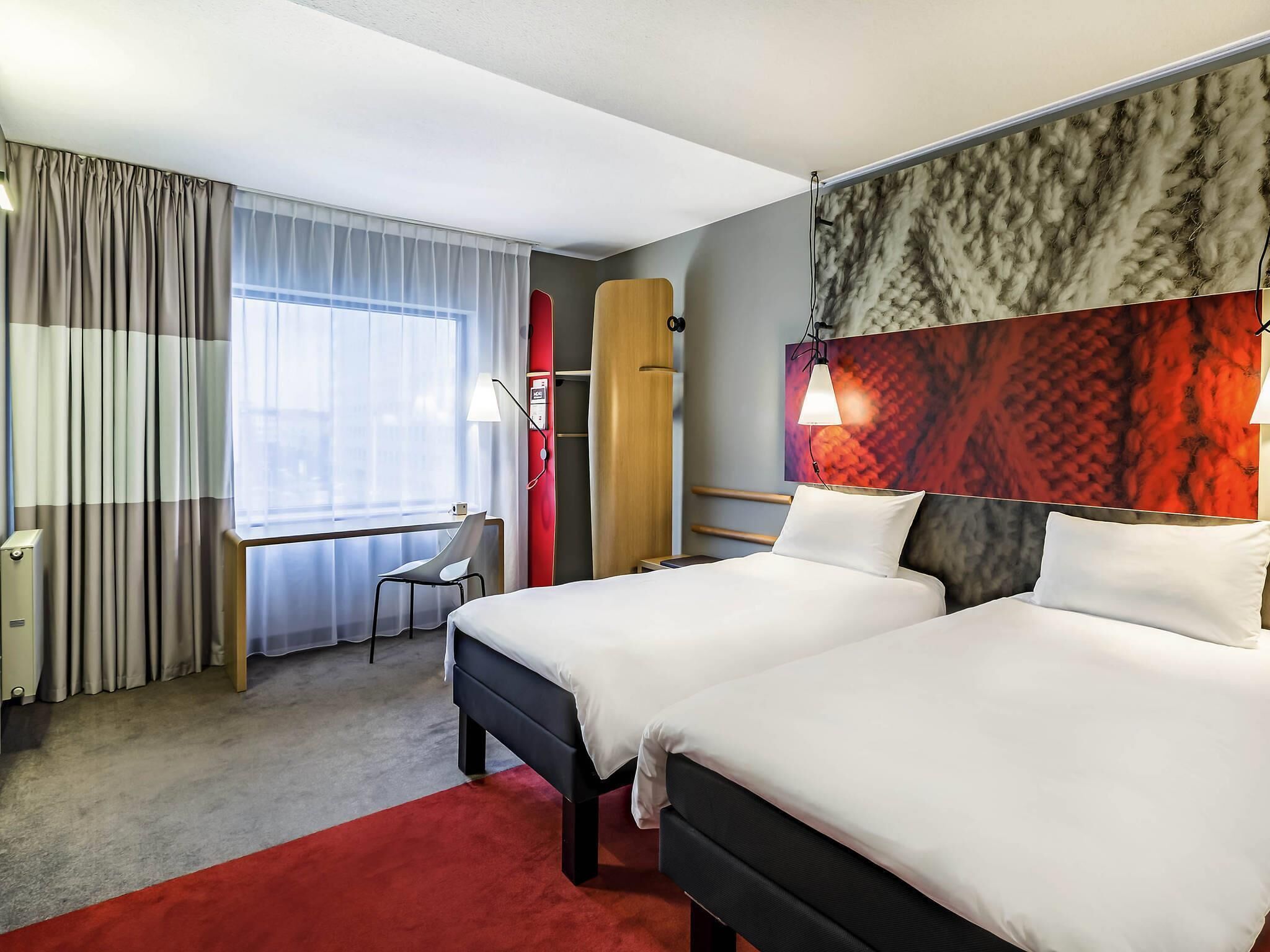 Una habitación de hotel con dos camas, un escritorio, una silla, una alfombra roja y una ventana con cortinas.