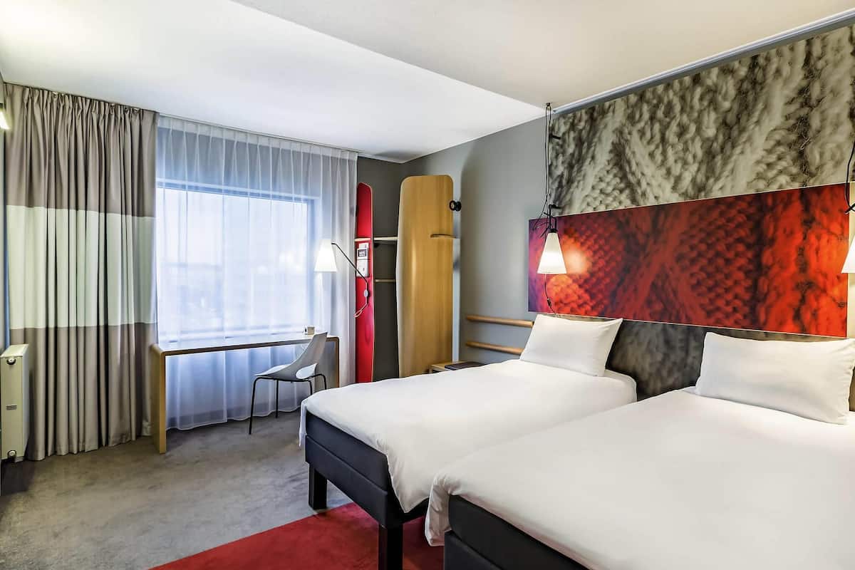 Una habitación de hotel con dos camas, un escritorio, una silla, una alfombra roja y una ventana con cortinas.
