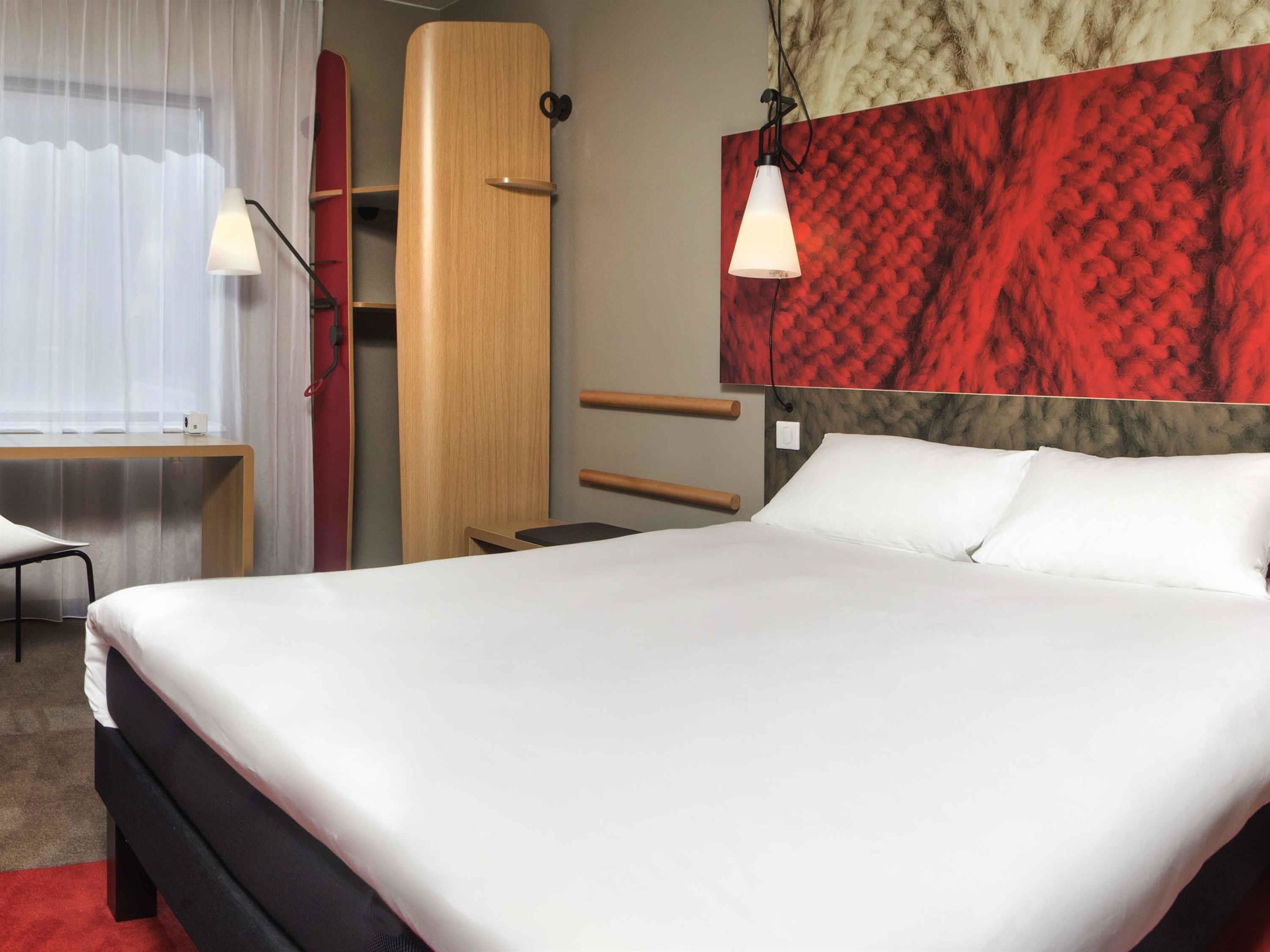 A hotel room with a large bed, a desk, a chair, a wardrobe, and a red textured wall.