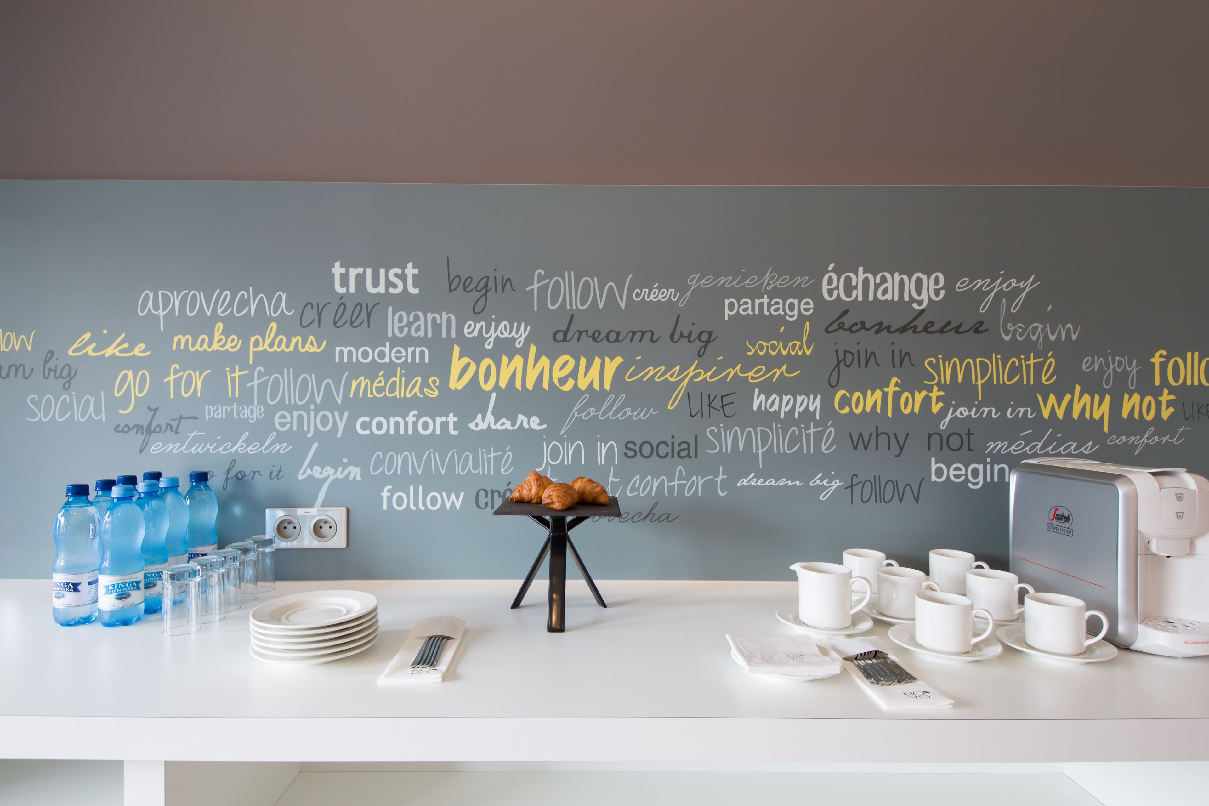 Una habitación moderna con una nube de palabras en la pared, una cafetera, tazas, platos y botellas de agua sobre la mesa.