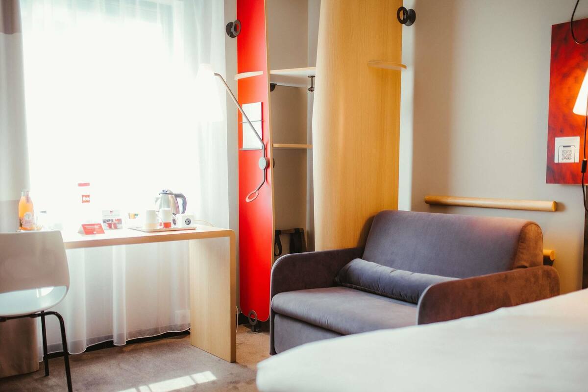 A hotel room with a bed, a desk with a chair, a wardrobe, and a bedside table with a lamp.