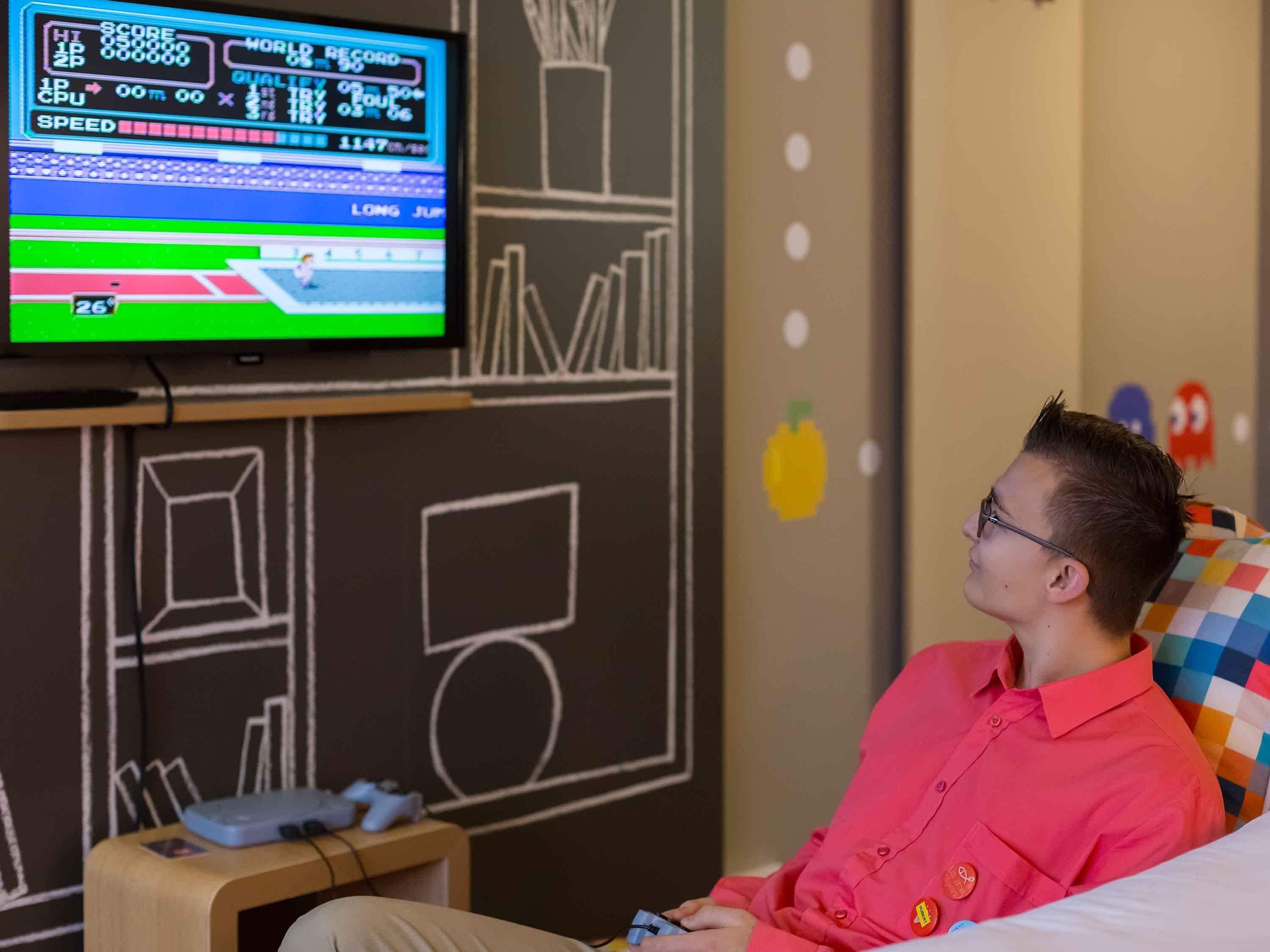 A person seated in a room with a television displaying a video game, a wall with chalk drawings, and a small table with items on it.