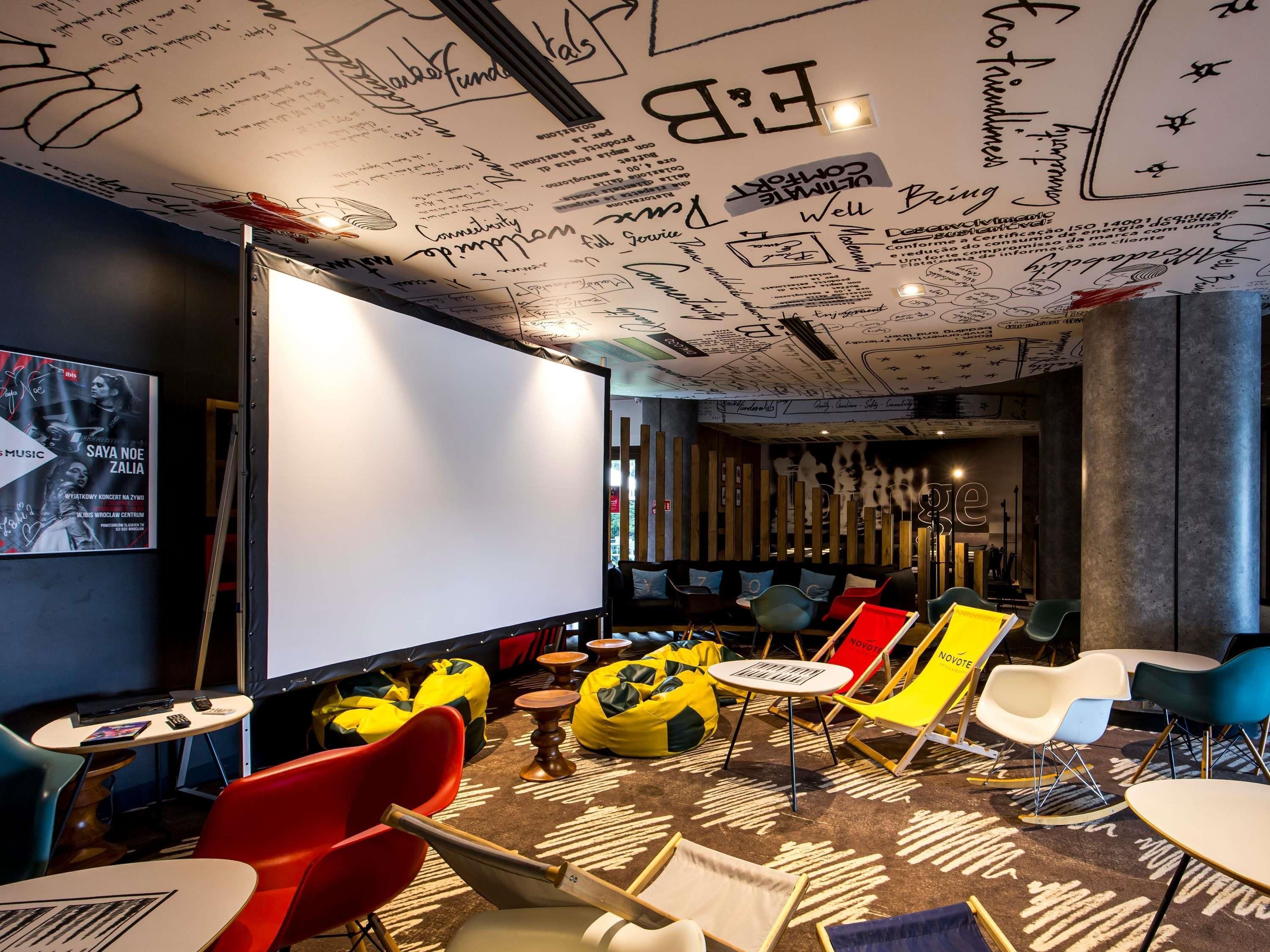 A modern lounge area with a large screen, colorful chairs, and a ceiling with artistic designs.
