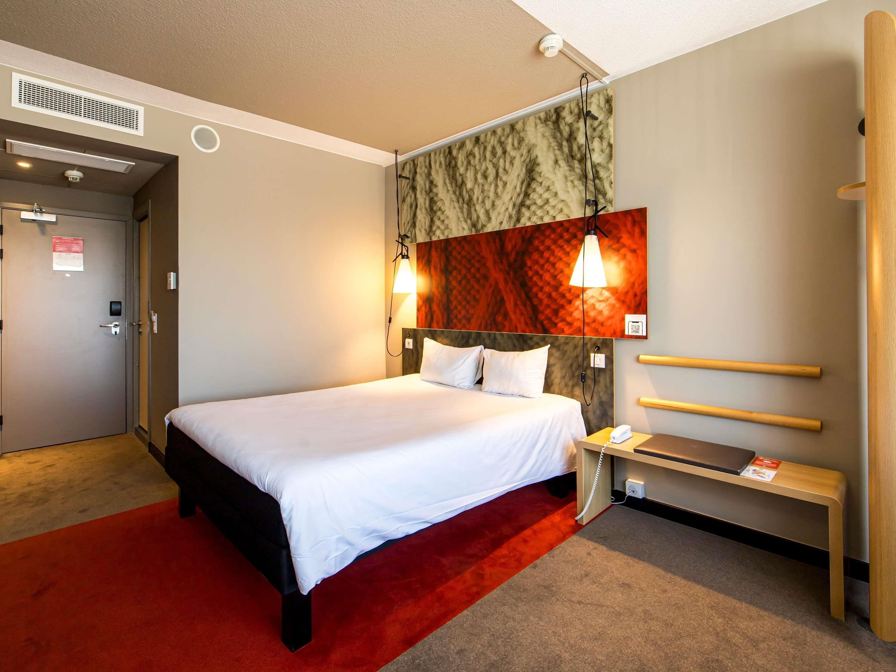 Een hotelkamer met een bed, een nachtkastje met een telefoon, een rood tapijt, een wandlamp en een deur met een beveiligingscamera.