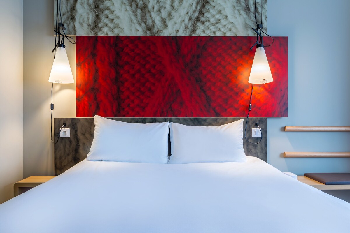 A neatly made bed with white linens, a red abstract wall art above, and two pendant lights hanging above.