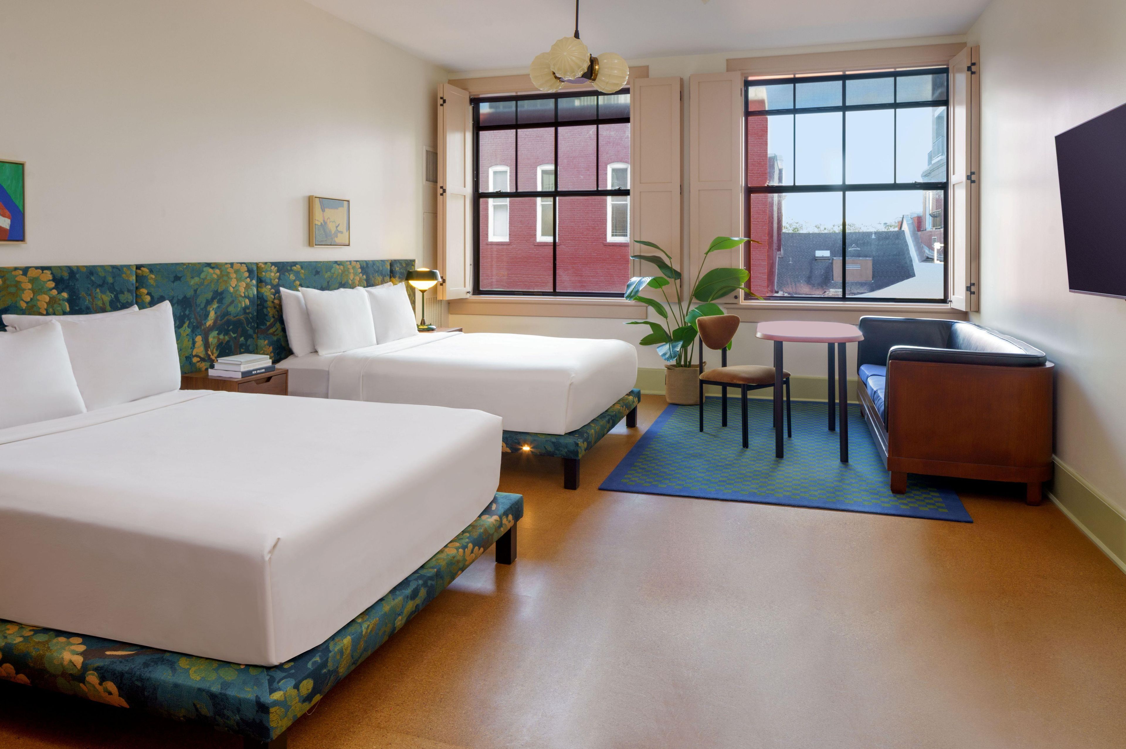 A hotel room with two beds, a desk, a chair, a small table, and a large window with a view of buildings.