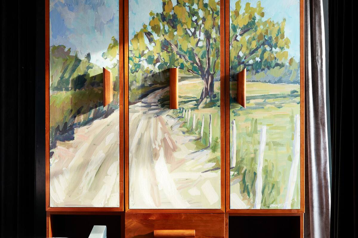 A room with a large painting of a tree-lined path, a wooden cabinet with drawers, and a bed with a patterned cover.