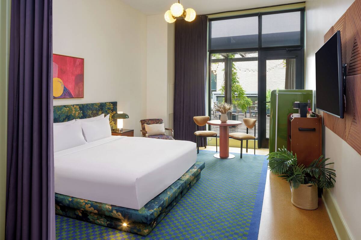 Hotelkamer met een groot bed, een bureau, een stoel, een kleine tafel, een televisie en een balkon met planten.
