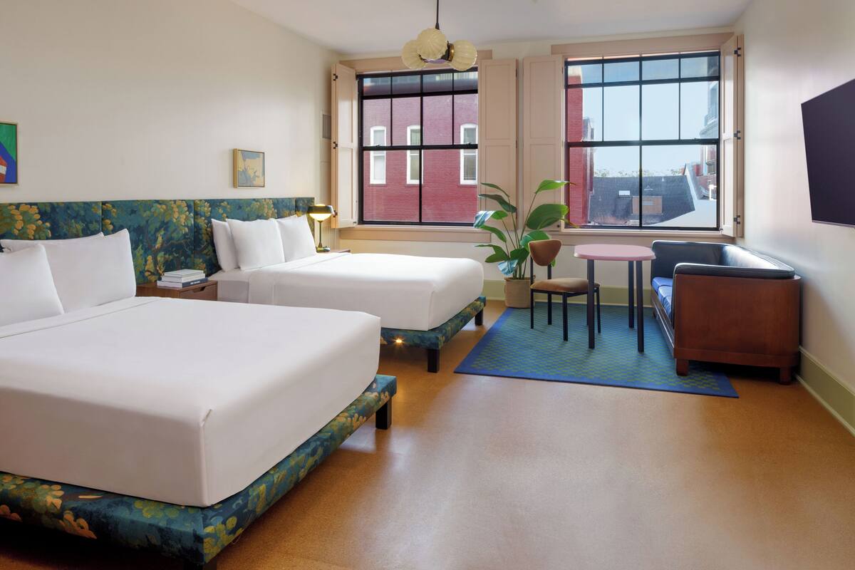 A hotel room with two beds, a small table, a chair, a plant, and a TV on the wall.