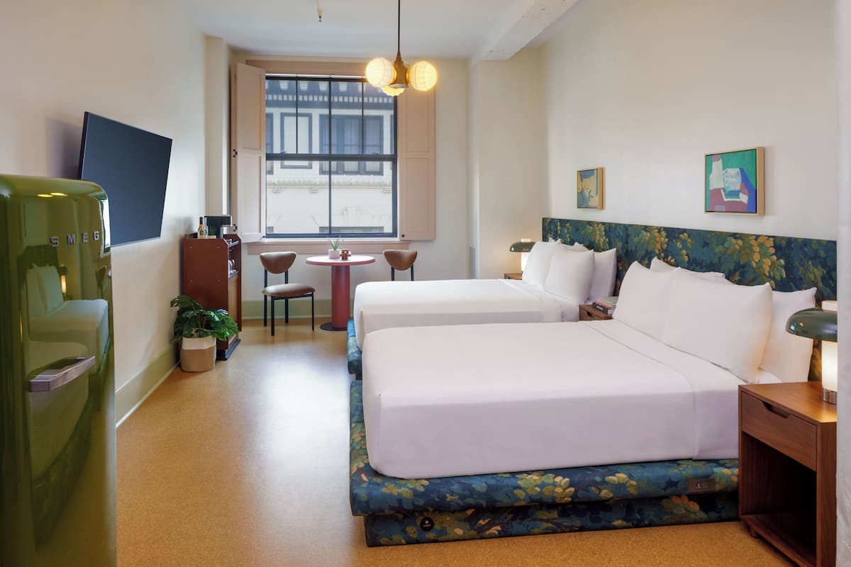 A hotel room with two beds, a small table, chairs, a TV, and a window with a view of buildings.