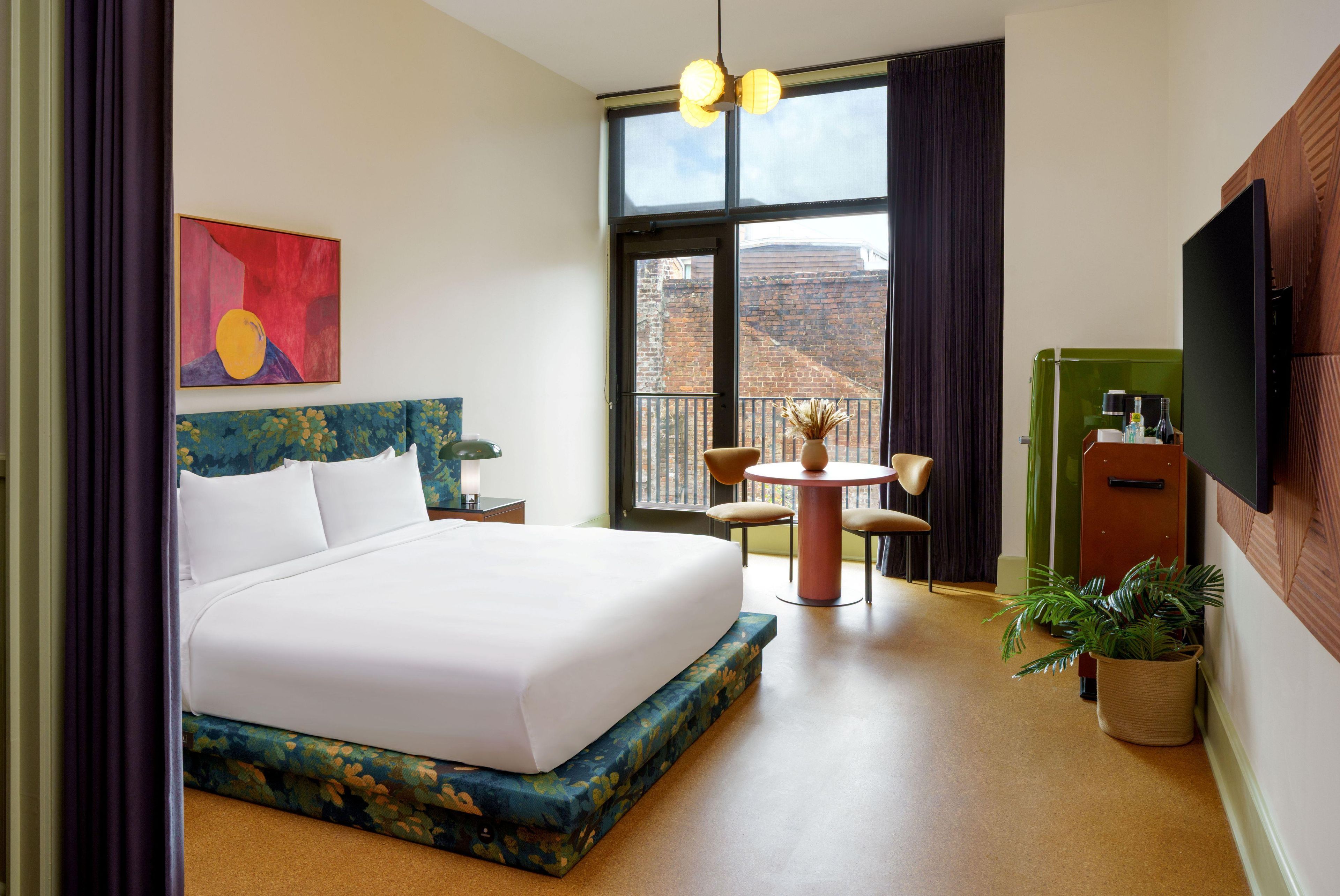 A hotel room with a large bed, a desk, a chair, a small table, a TV, and a view of the outdoors.