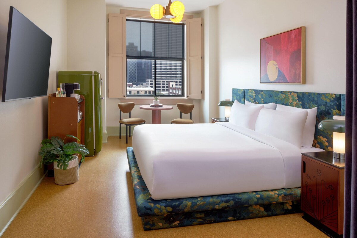 Hotelkamer met een groot bed, een bureau met lamp, een televisie, een raam met uitzicht op de stad en een plant.