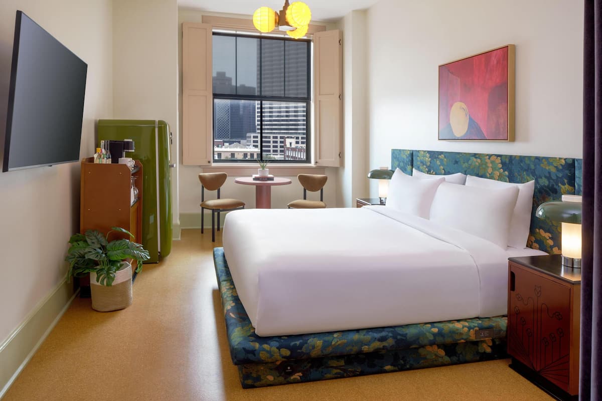 A hotel room with a large bed, a desk with a lamp, a television, a window with a city view, and a plant.