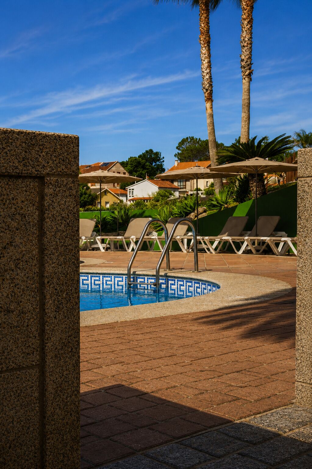 Zona de piscina con sillas de descanso, piscina con mosaicos azules y palmeras.