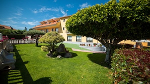 A well-maintained garden with a variety of plants and a large tree, a building with a tiled roof, and a clear blue sky. - Hotel Atlántico Sanxenxo (Sanxenxo)
