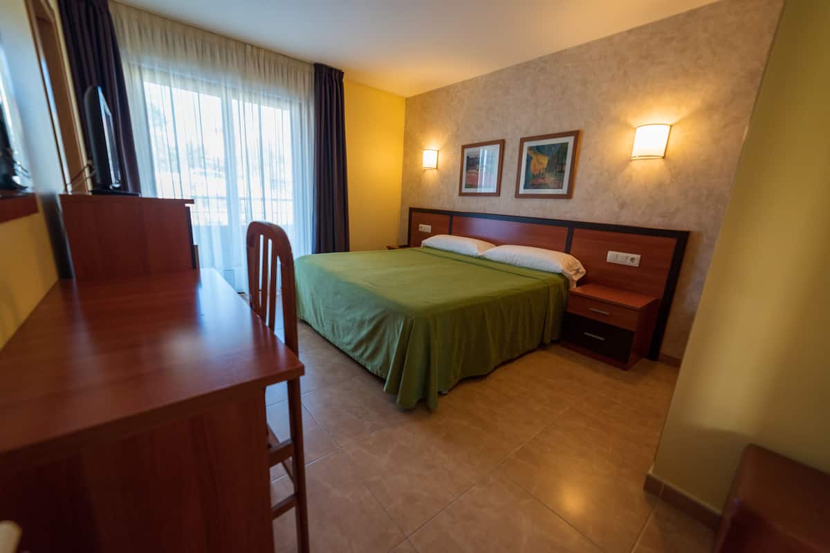 A hotel room with a bed, a desk, a chair, a nightstand, and two wall-mounted lamps.
