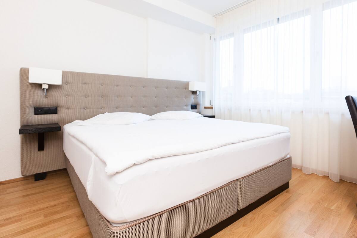Ein ordentlich bezogenes Bett in einem Hotelzimmer, mit Kopfteil, Nachttisch und einem Sessel.