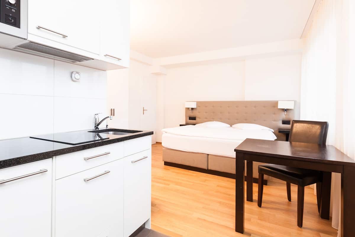 Ein modernes Hotelzimmer mit Küchenzeile, einem Bett mit weißer Bettwäsche, einem Holztisch und einem Nachttisch mit Lampe.