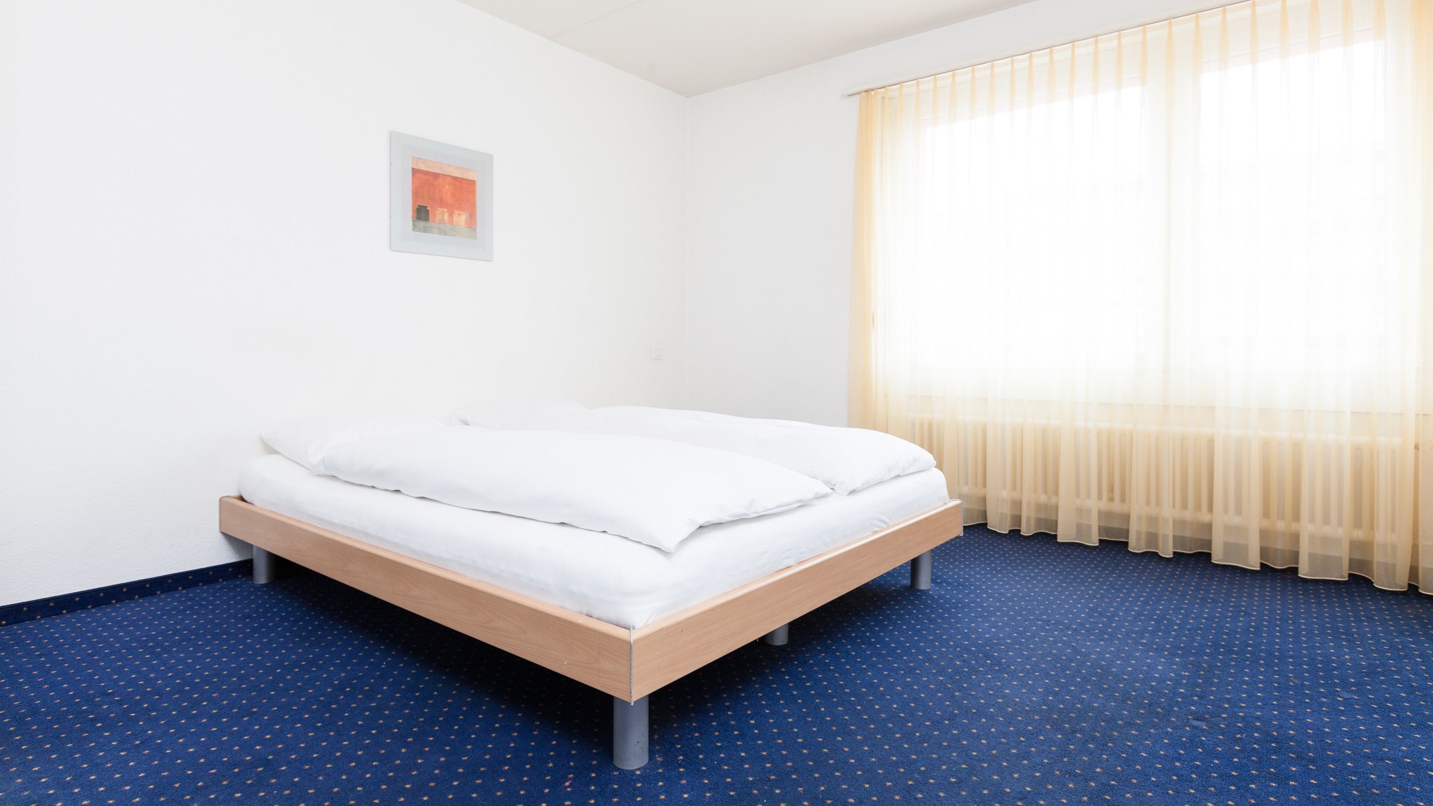 Ein Einzelbett in einem Zimmer mit blauem Teppich, weißen Wänden und einem Fenster mit durchsichtigen Vorhängen.