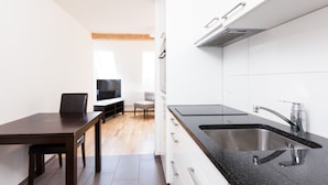 Ett modernt kök med en svart bänkskiva, en diskho i rostfritt stål och vita köksskåp. Det finns en matplats med ett mörkt träbord och stolar, och en tv i bakgrunden.