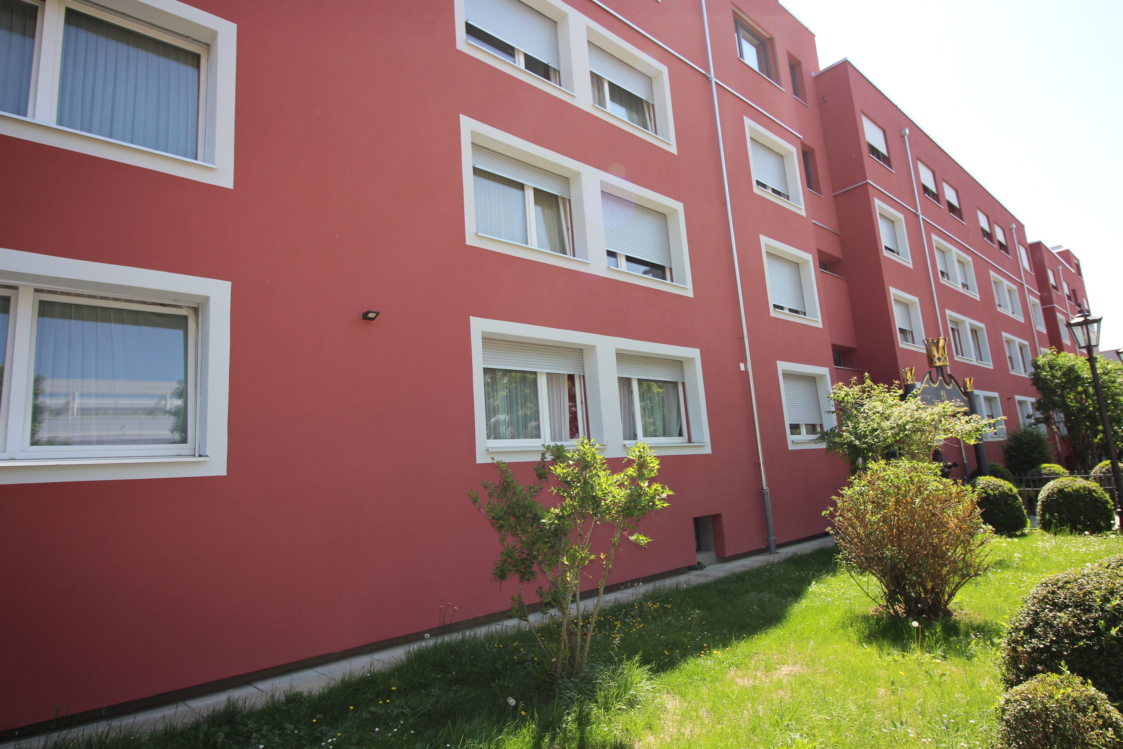 A red building with multiple windows and a small garden area with trimmed bushes.