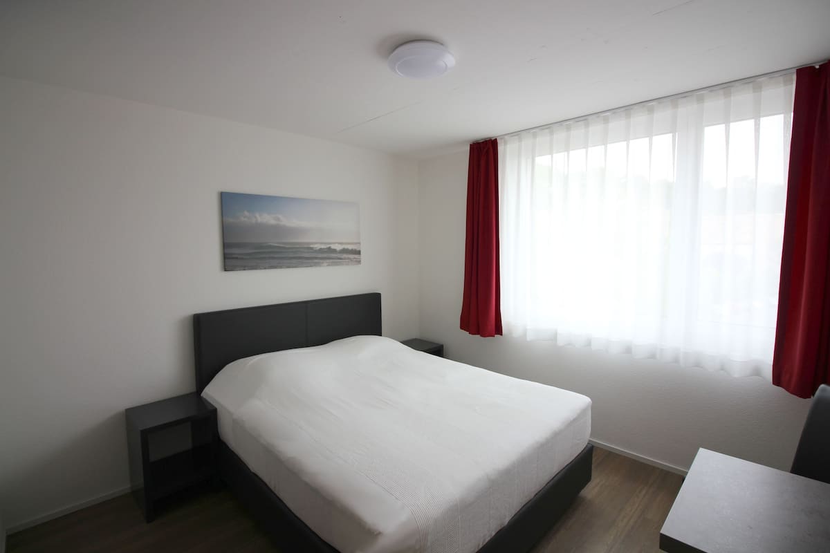 Ein Hotelzimmer mit Bett, Nachttisch, kleinem Tisch und einem Fenster mit Vorhängen.