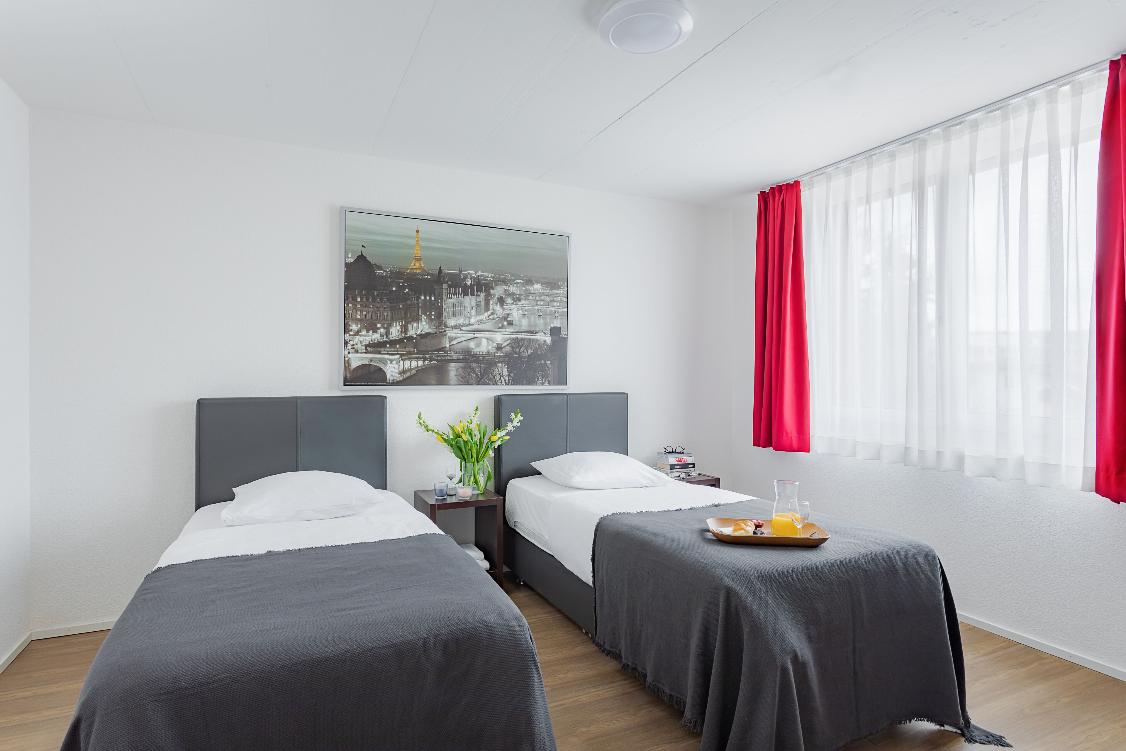 A hotel room with two beds, a nightstand, a small table with a tray of breakfast, a window with red curtains, and a cityscape painting on the wall.