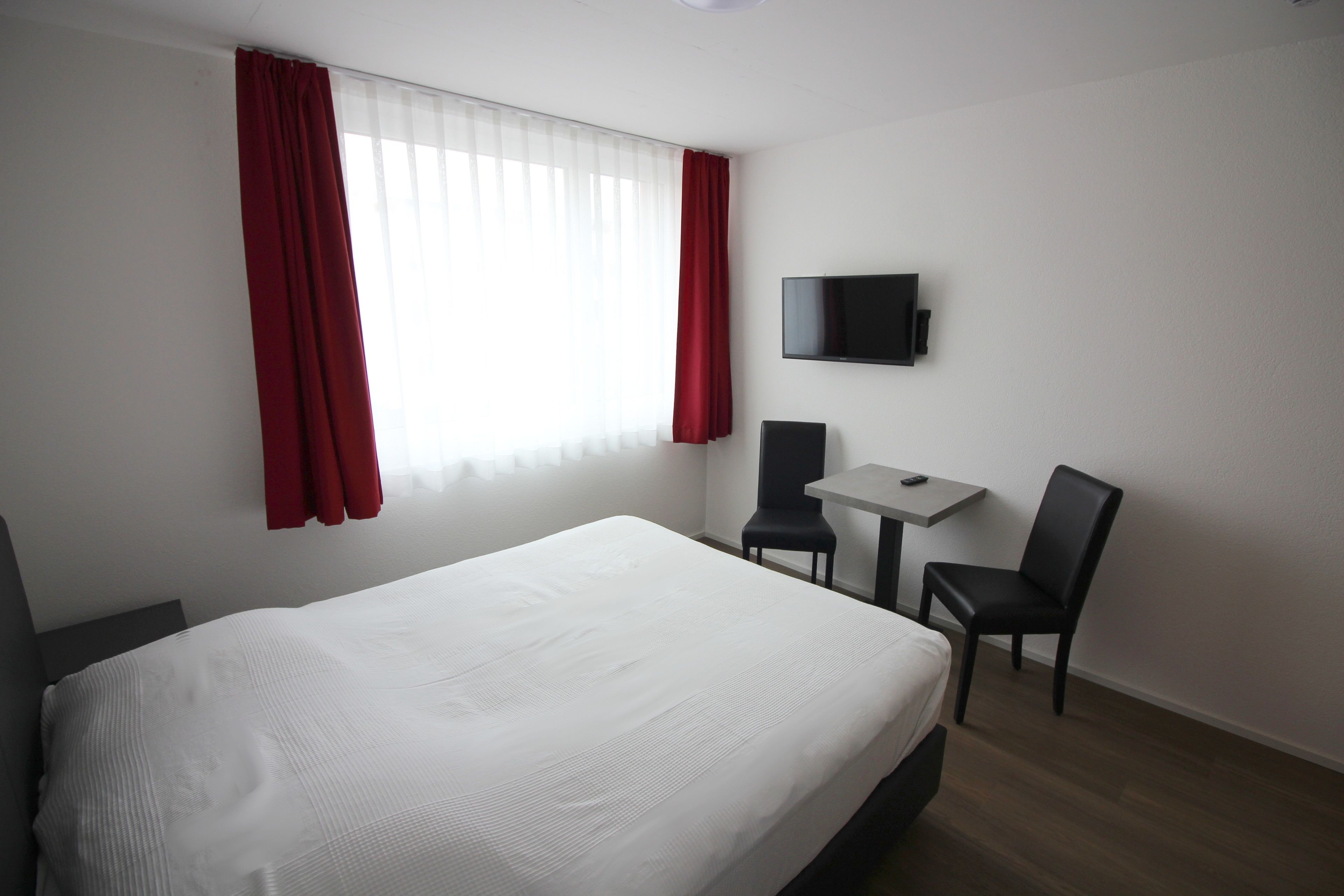 A hotel room with a large bed, a small table, two chairs, a TV mounted on the wall, and a window with red curtains.