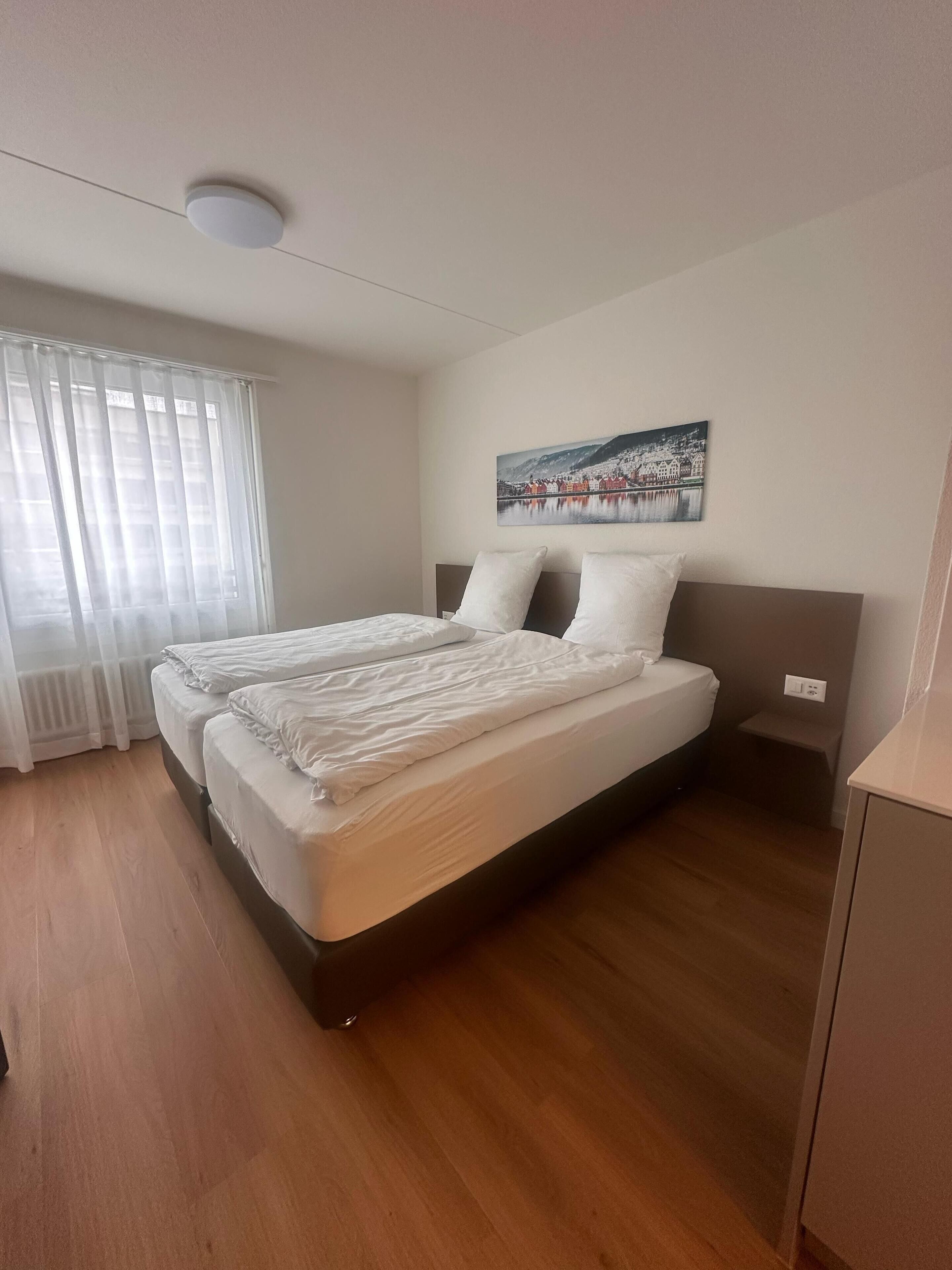 Un llit doble amb roba de llit blanca en una habitació amb terra de fusta, una làmpada al sostre, una finestra amb cortines translúcides i un quadre penjat a la paret.