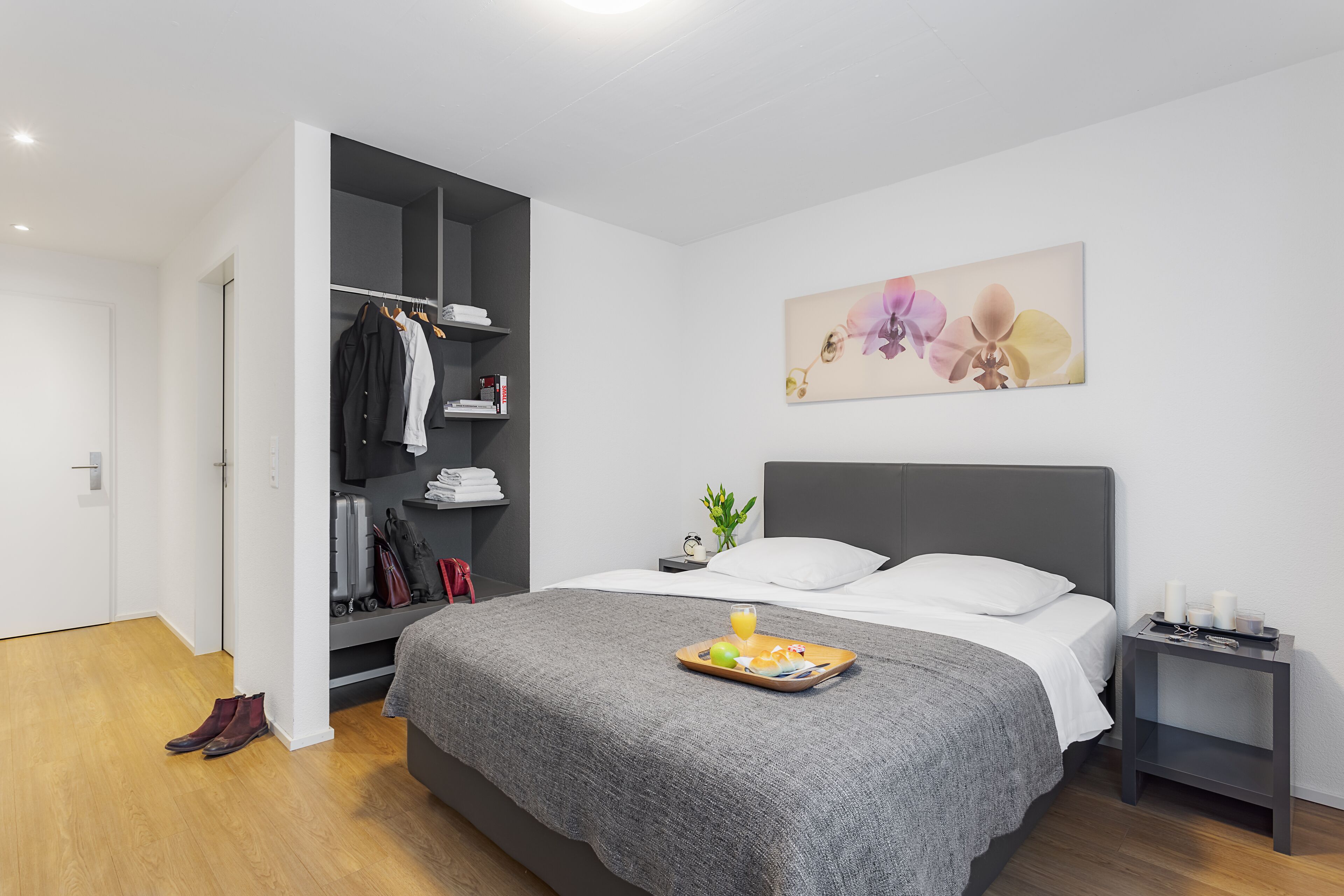 Ein modernes Schlafzimmer mit einem grauen Bett, einem Nachttisch mit Frühstücksbrettchen, einem Kleiderschrank und einem an der Wand befestigten Orchideenbild.