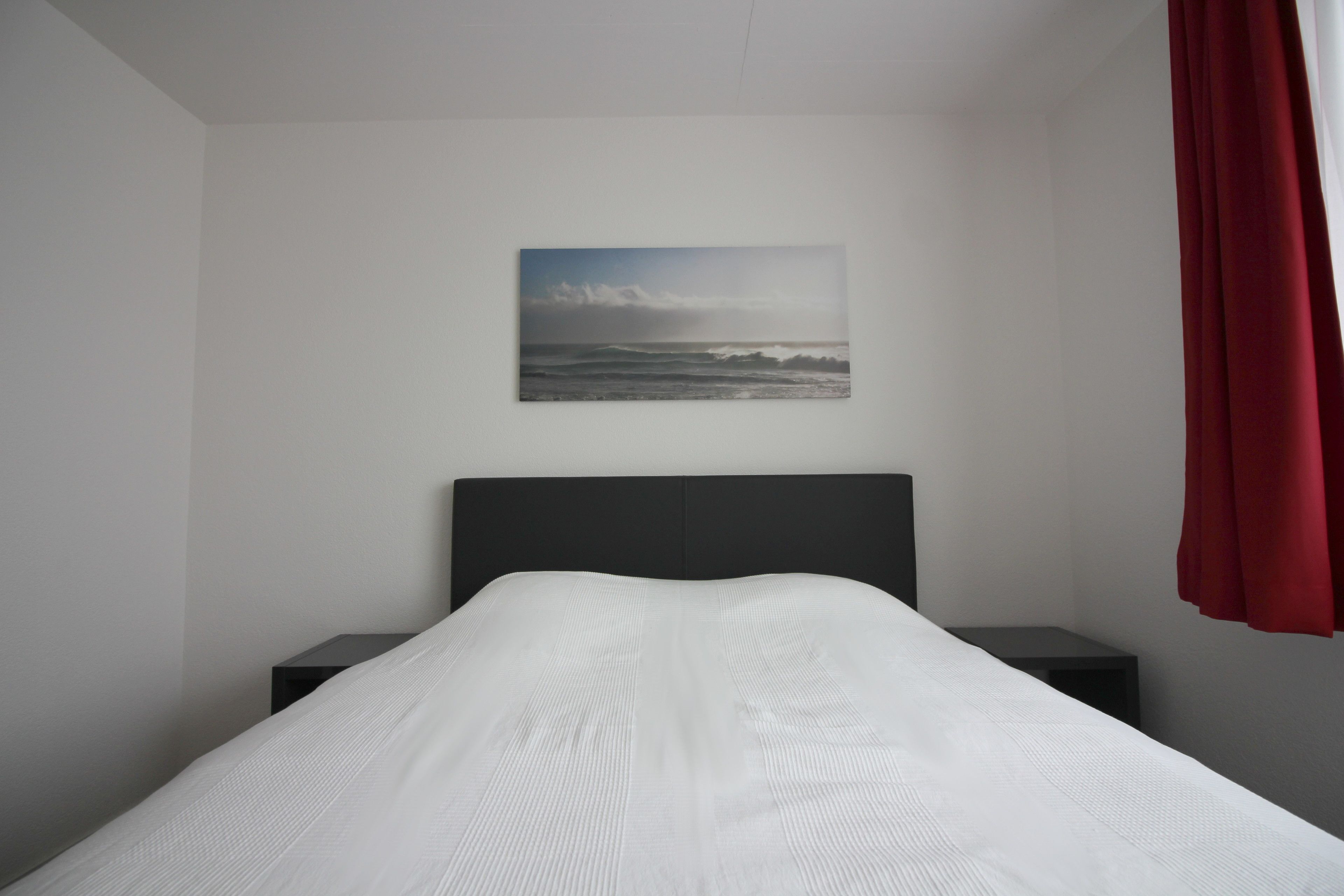 A bedroom with a bed, a nightstand, a wall-mounted picture, and a red curtain.