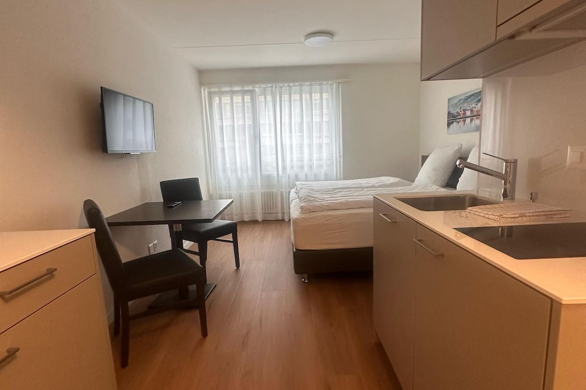Ein kleines Hotelzimmer mit Bett, Schreibtisch, Stuhl, Wand-Fernseher, einem Fenster mit leichtem Vorhang und einer Küchenzeile mit Spüle und Herd.