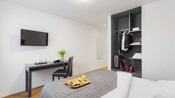 A modern hotel room with a bed, a desk with a chair, a flat-screen TV, a wardrobe, and a tray with breakfast items.