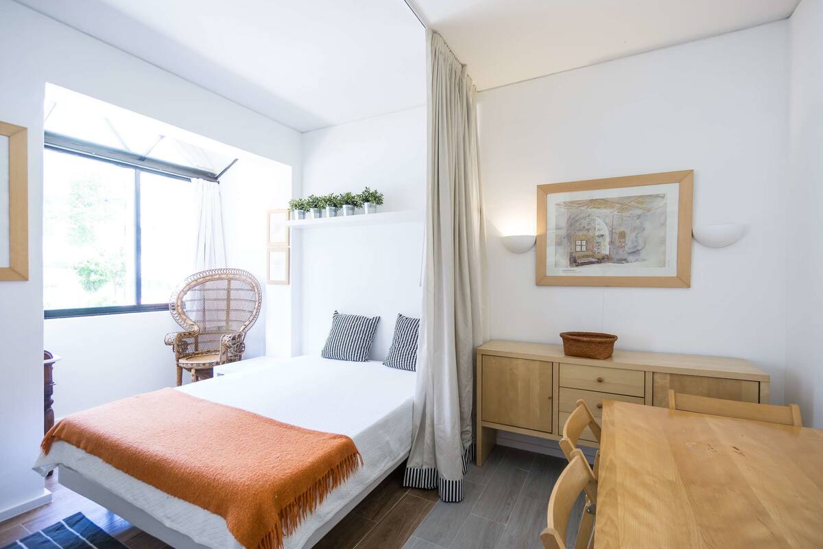 A bedroom with a bed, a chair, a wooden table, a cabinet, and a window with curtains.