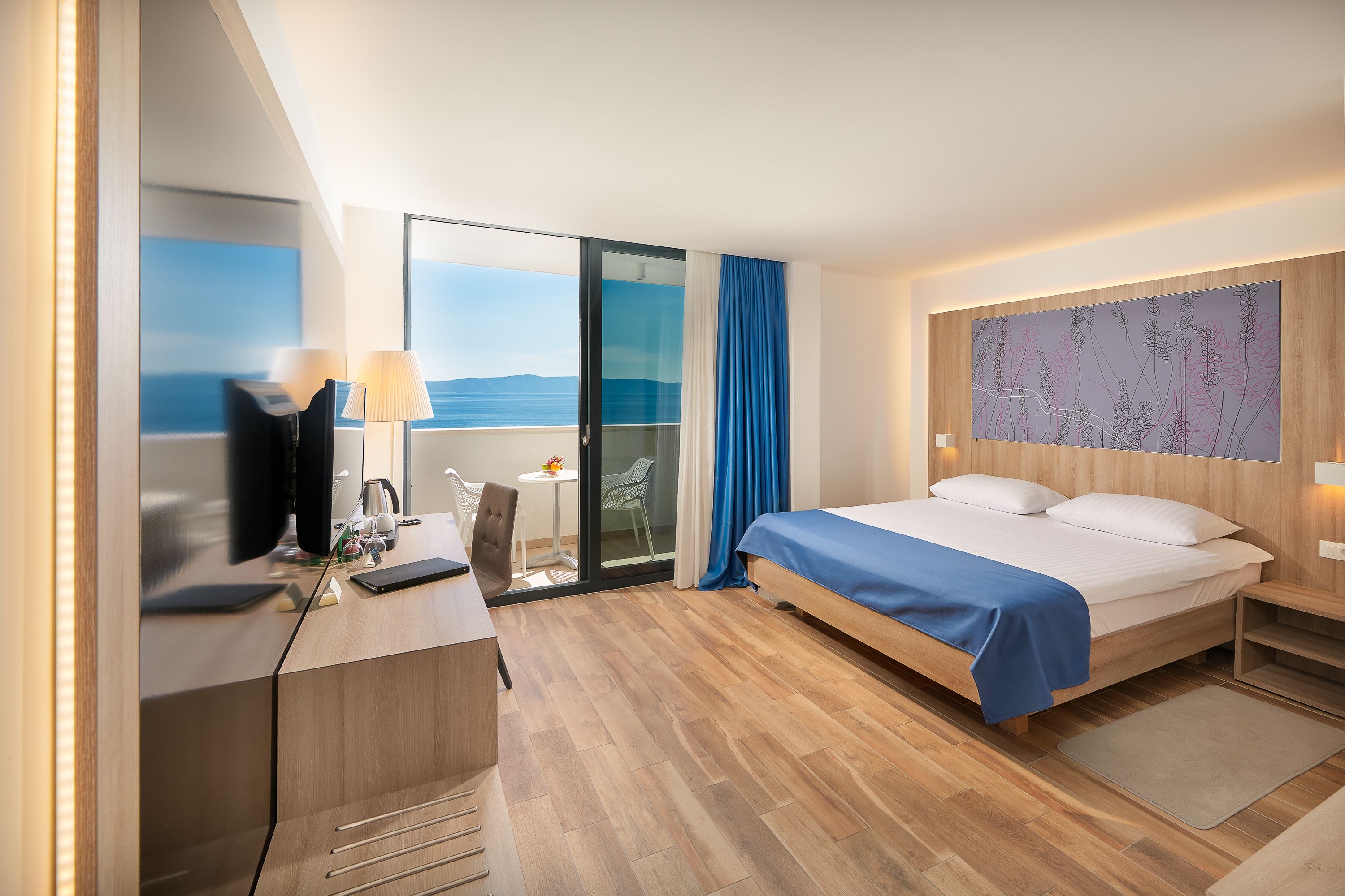 A modern hotel room with a bed, desk, and a view of the sea.