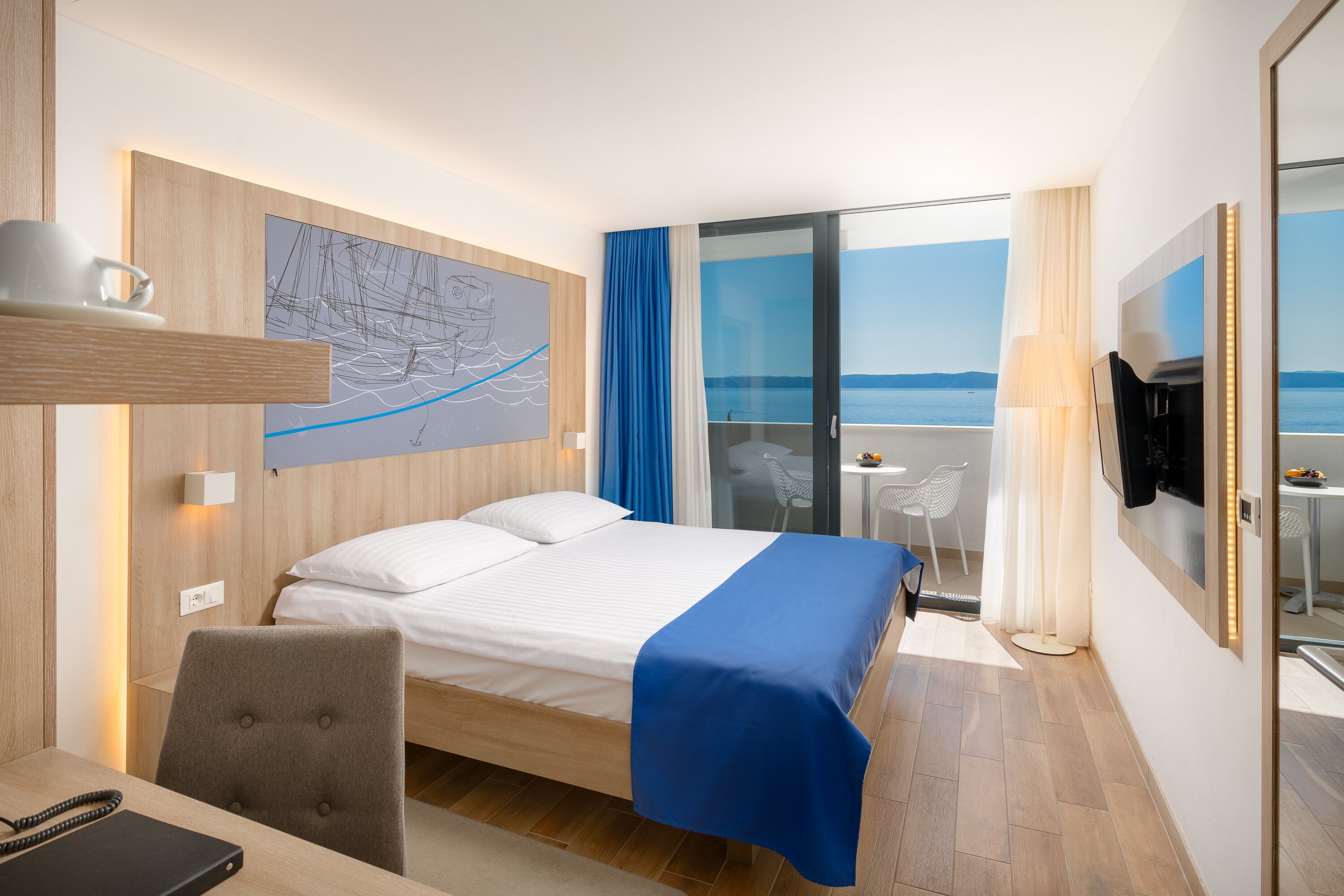 A hotel room with a bed, a desk, a chair, a TV, a window with a view of the sea, and a painting on the wall.