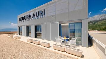 Un edificio moderno con el nombre 'MEDORA AURI' en la fachada, mobiliario de mimbre en la terraza exterior y vista al mar.