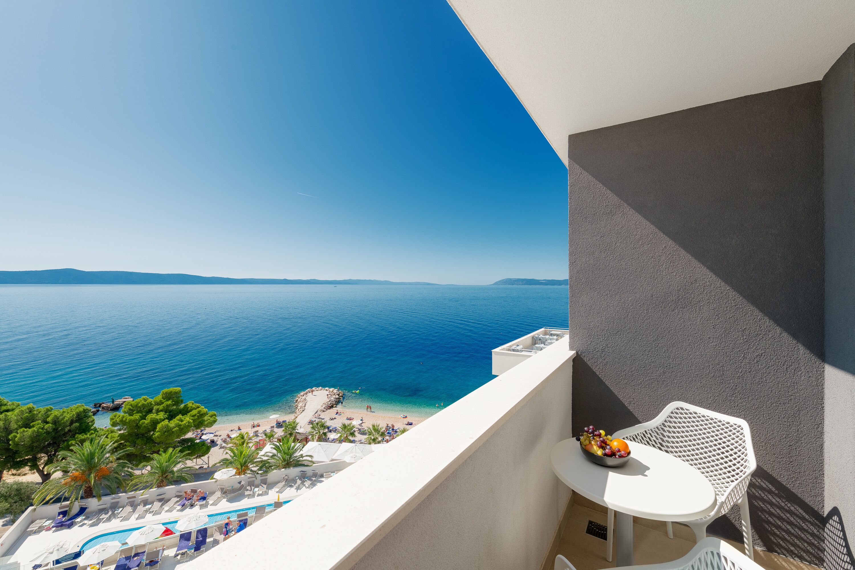 A balcony with a view of the sea, a white table with a fruit bowl, and a pool area with lounge chairs.