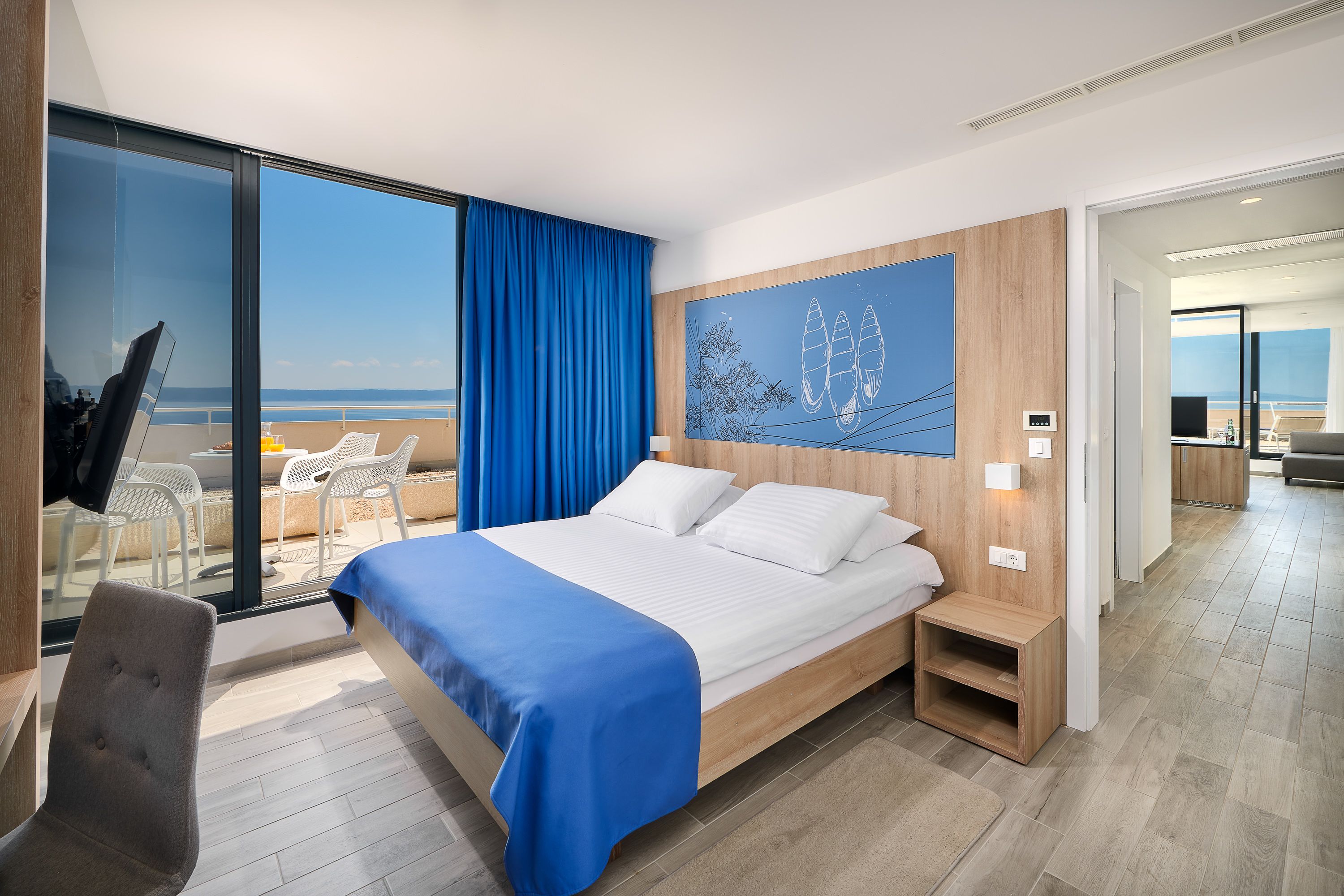 A modern hotel room with a large bed, a view of the beach, and a dining area visible through an open door.