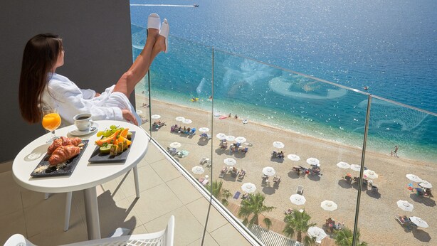 A balcony with a table set for breakfast, overlooking a beach with umbrellas and people.